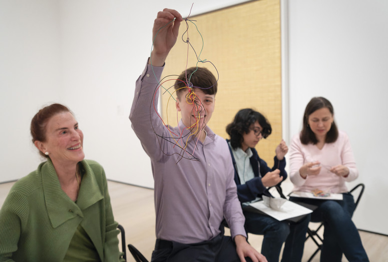 Photo: Martin Seck. © 2024 The Museum of Modern Art, NY Image Description: A smiling man wearing a purple button-up shirt holds up a sculpture of tangled colorful wires all tied together. His mother wearing a green cardigan smiles up at his artwork in the blurred background.
