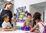 Photo: onwhitewall.com. © 2024 The Museum of Modern Art, NY Image Description: Three children draw at a table covered with coloring paper and a purple box of markers while a red-haired woman in a black dress looks over them and smiles.