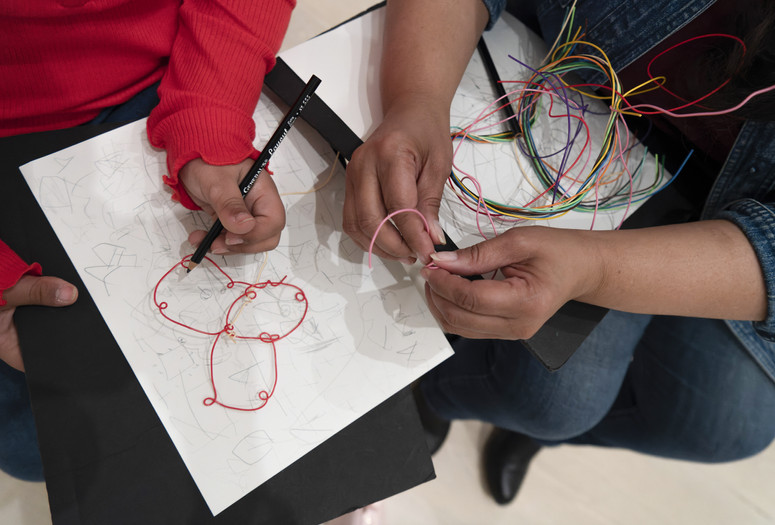 Photo: Martin Seck. © 2023 The Museum of Modern Art, NY Image Description: The hands of a girl with red shirt sleeves draw with a black pencil on a white piece of paper. A red bent wire sculpture sits on the page and the hands of her mother fold a bright pink wire on top of a bunch of colorful wires in her lap.