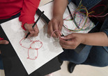 Photo: Martin Seck. © 2023 The Museum of Modern Art, NY Image Description: The hands of a girl with red shirt sleeves draw with a black pencil on a white piece of paper. A red bent wire sculpture sits on the page and the hands of her mother fold a bright pink wire on top of a bunch of colorful wires in her lap.