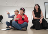 Photo: Martin Seck. © 2023 The Museum of Modern Art, New York Image description: A young woman wearing a coral pink shirt and pink headband sits cross-legged on a gallery floor between her mother, pointing off to the left, and a crouched educator in a long black dress looking up in the same direction.