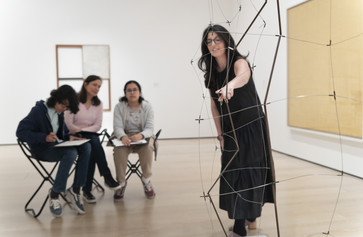 Photo: Martin Seck. © 2023 The Museum of Modern Art, NY Image Description: An educator in a black dress points to a hanging wire sculpture by the artist Gego, while a family of three people sitting on gallery stools look at the artwork and draw together.