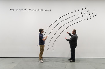 Photo: Martin Seck Image description: Two men sign to each other while standing in front of a black-and-white line drawing mural by the artist Christine Sun Kim. &#34;The Sound of Temperature Rising&#34; is written in capital letters at the top left corner, while the men stand in front of four arching lines that mimic music stanzas.
