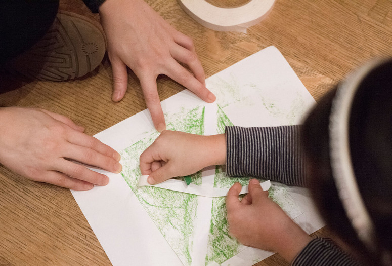 Photo: Martin Seck Image description: Adult hands with white skin hold down a piece of white paper on the floor. The paper has a square made of light green crayon. A child leaning over the paper is arranging a white letter "X" out of two strips of paper onto the the green square.