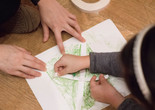 Photo: Martin Seck Image description: Adult hands with white skin hold down a piece of white paper on the floor. The paper has a square made of light green crayon. A child leaning over the paper is arranging a white letter &#34;X&#34; out of two strips of paper onto the the green square.