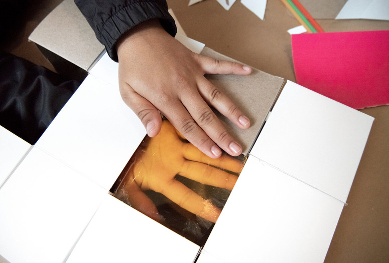 Photo: Martin Seck Image description: A child's hand with brown skin is holding a cardboard rectangle with a transparent orange square in the center. The child's other hand can be seen, face up, pressed against the underside of the orange square.