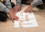 Photo: Martin Seck Image description: The hands of a child and adults with brown skin arrange rectangular pieces of cut paper with yellow, blue, and red squares on them on a white piece of paper on the floor.