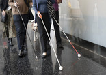 Photo: Martin Seck Image description: Cropped photo of three people walking with navigation canes down a hallway with a black marbled floor and a reflective glass wall. Each cane is different—the leftmost is black with a small tip, the middle is white with a large white ball tip and an orange handle, and the rightmost is white and red with a large white ball tip. Behind the group are a few more people and a pale yellow dog in a harness.