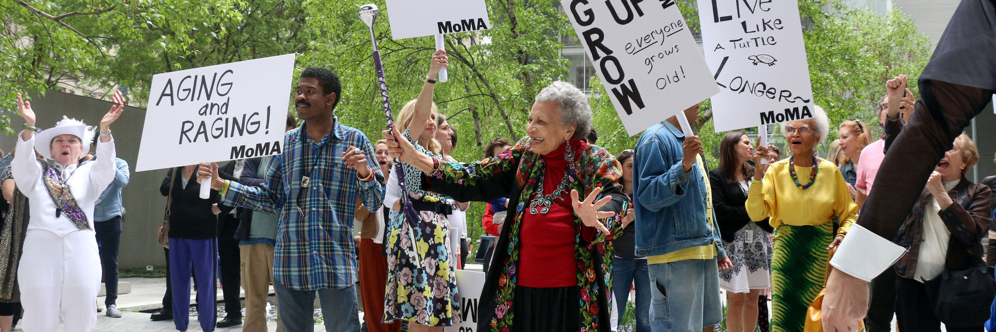 Photo: Manuel Molina Martagon. ©️ 2023 The Museum of Modern Art, New York Image description: A group of older adults are gathered outside in MoMA’s Sculpture Garden with a marching band. The crowd wears colorful clothing and hold signs that read, “Aging and Raging!” and “Grow Up! Everyone Grows Old!”