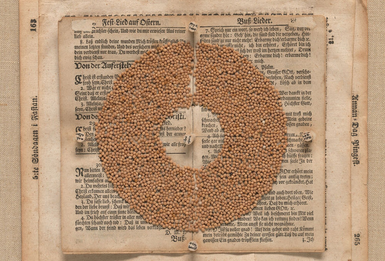 Lenore Tawney. Seed Circle. 1967. Ink on corks and seeds on book pages. The Museum of Modern Art, New York. Gift of Sarah-Ann and Werner H. Kramarsky. © 2023 Lenore Tawney Image description: Beige circular seeds lay in a thick ring, barely divided in half by the binding of the open pages they rest on. Discolored and weathered book pages display medieval calligraphy in dark ink. Five corks, one in the center and four others on the top, bottom, left and right of the circle of seeds, have small notes written in black.