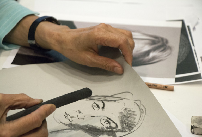 Photo: Manuel Molina Martagon Image description: In a close-up photograph of an older woman’s hands, she holds a piece of charcoal in her right hand and paper with a charcoal drawing of a woman in her left.