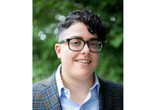 Aimi Hamraie Image description: Aimi Hamraie, an olive-skinned Iranian person with short dark curly hair, smiles at the camera. They wear rectangular glasses and a blue button-up shirt under a blue-green checkered blazer. Behind them is a blurred green background.