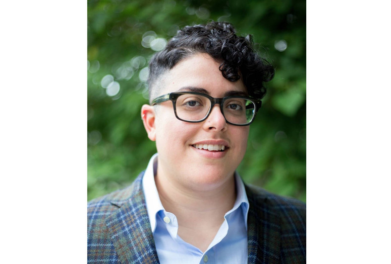 Aimi Hamraie Image description: Aimi Hamraie, an olive-skinned Iranian person with short dark curly hair, smiles at the camera. They wear rectangular glasses and a blue button-up shirt under a blue-green checkered blazer. Behind them is a blurred green background.