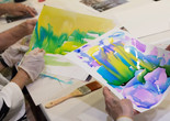 Photo: Manuel Molina Martagon. © 2024 The Museum of Modern Art, New York Image description: A photograph showing the hands of older adults holding watercolor paintings. On the right, the hands hold a wet composition of melding purple, blue, and yellow. To the left, gloved hands paint broad yellow strokes. A wet brush sits on the table between them.