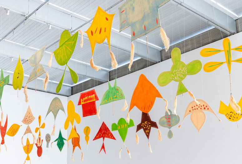 Installation view of Joan Jonas: Good Night Good Morning, The Museum of Modern Art, New York, March 17–July 6, 2024. Photo: Jonathan Dorado. Digital image © 2024 The Museum of Modern Art, New York Image description: An installation of 30 colorful paper kites hanging in three rows from the rafters of a white industrial ceiling. The shapes of the kites are different amorphous forms. They are hand-painted in brilliant yellows, greens, oranges, and reds, with a few brushstrokes in contrasting colors dotting each surface. Some have tufts of tan raffia drifting down from their corners.