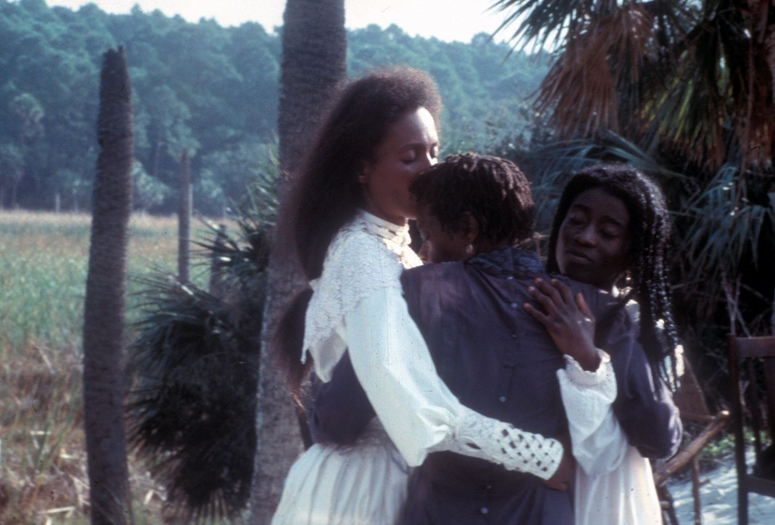 Daughters of the Dust. 1991. USA. Directed by Julie Dash. The Museum of Modern Art, New York. Film Study Collection Image Description: Three Black women embrace with pensive, furrowed expressions. They stand in a tropical field, surrounded by palm trees and tall grasses. A lush forest stands in the hazy background.