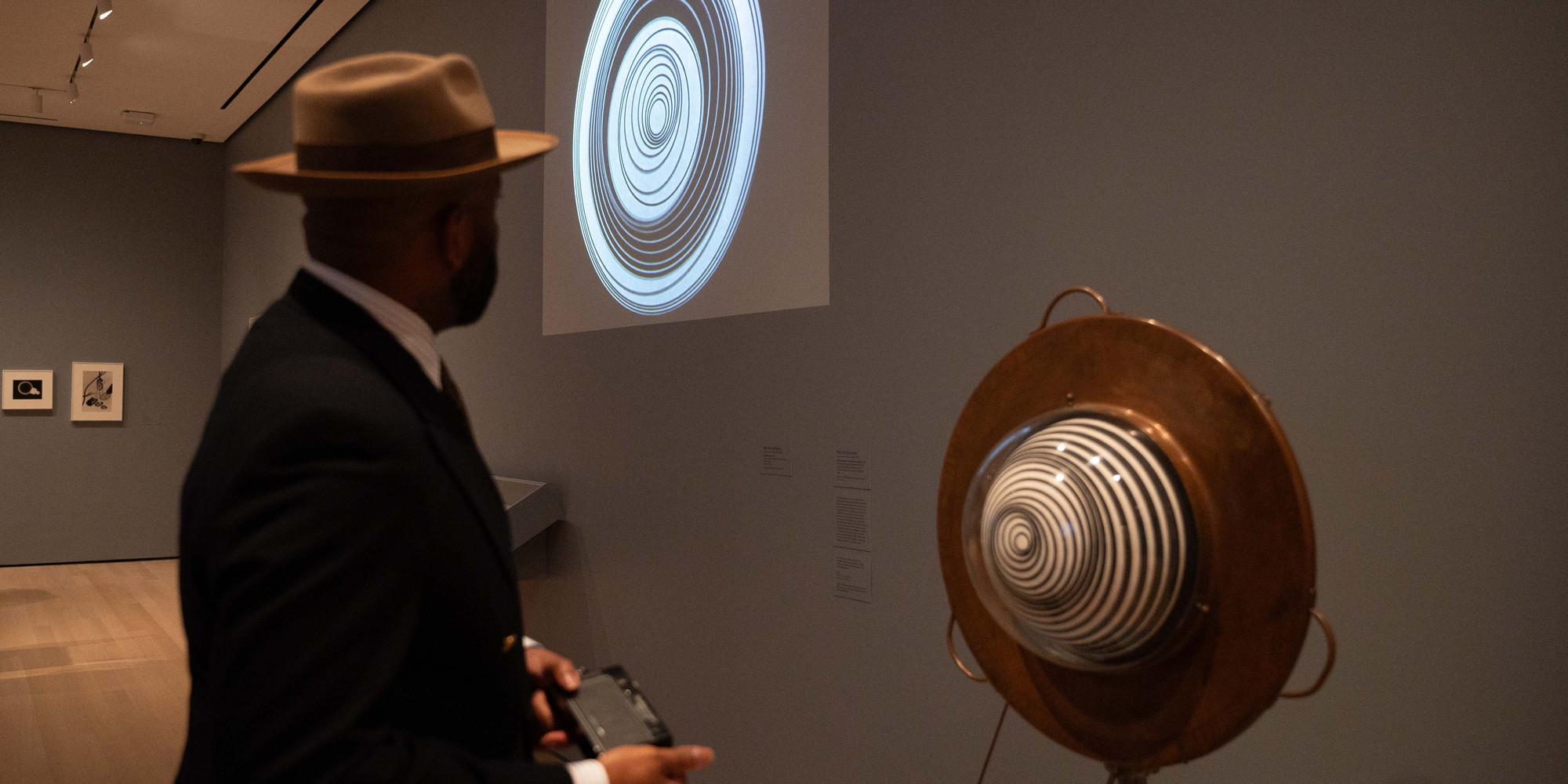 Naeem Douglas at MoMA. Photo: Natiah Jones. Shown: Marcel Duchamp Rotary Demisphere (Precision Optics). 1926. 35mm film transferred to video, 6 min. at 22 fps. The Museum of Modern Art, New York, 1938. Anemic Cinema. Paris, 1925. Painted papier-mâché demisphere fitted on velvet-covered disk, copper collar with plexiglass dome, motor, pulley, and metal stand, 58 1/2 × 25 1/4 × 24" (148.6 × 64.2 × 60.9 cm). The Museum of Modern Art, New York. Gift of Mrs. William Sisler and Edward James Fund. © 2024 Artists Rights Society (ARS), New York/ADAGP, Paris/Estate of Marcel Duchamp