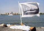 LaToya Ruby Frazier. Holding flag laying at the edge of Pier 54 and the Hudson River. On flag: US Army transport Buford, the “Soviet ark,” used to deport political radicals and other “undesirable” noncitizens from the United States to Russia in December 1919, broadside view, 1907 from Pier 54: A Human Right to Passage, 2014. © 2024 LaToya Ruby Frazier. courtesy of the artist and Gladstone gallery