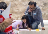 Photo: Beatriz Meseguer Image description: A child and father sit on the floor looking down at their drawing in front of the painting Houston Street by Martin Wong