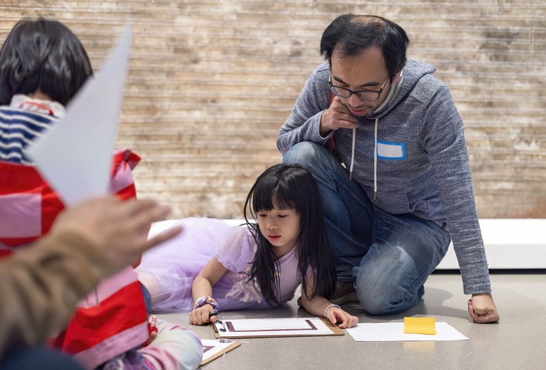 Photo: Beatriz Meseguer Image description: A child and father sit on the floor looking down at their drawing in front of the painting Houston Street by Martin Wong