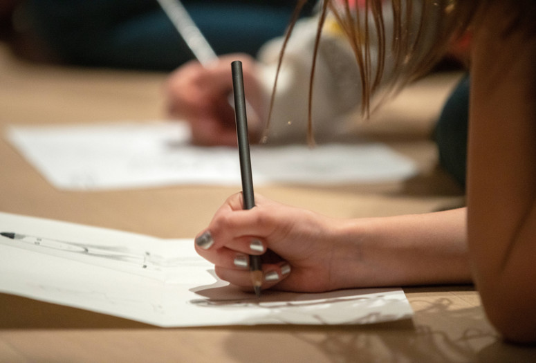 Photo: Manuel Martagon Image description: A close-up of a child’s hand holding a pencil and drawing on paper while sitting on the ground