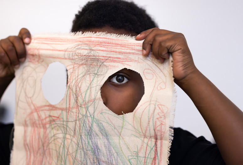 Photo: On White WallImage description: A child looks through a hole in their canvas drawing.