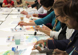 Photo: Manuel Molina MartagonImage description: Profile view of a long table of older adults painting with watercolors