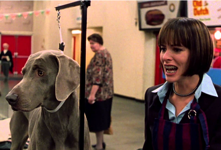 Best in Show. 2000. USA. Directed by Christopher GuestImage description: Film still of a Weimaraner dog secured by a leash on a grooming table next to a woman with a short bob of black hair, her mouth full of braces and agape in frustration.