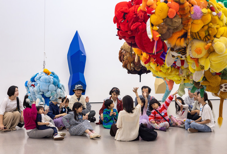 Photo: On White WallImage description: Children in a tour group raise their hands while sitting underneath floating sculptures.