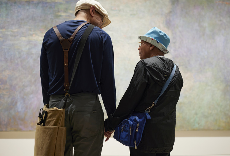 Photo: Jason LindbergImage description: Man faces away from the viewer, looking down to the right, and holds hands with his mother. His mother, a head shorter than him, looks in his direction with a slight smile. They stand in front of Claude Monet’s painting Water Lilies.