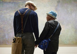 Photo: Jason LindbergImage description: Man faces away from the viewer, looking down to the right, and holds hands with his mother. His mother, a head shorter than him, looks in his direction with a slight smile. They stand in front of Claude Monet’s painting Water Lilies.