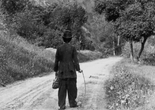 The Tramp. 1915. USA. Written and directed by Charles ChaplinImage description: Black and white film still of a person, with their back to the viewer, wearing a baggy suit and rounded top hat walking up an unpaved road through a woodsy landscape. They hold a long stick as a walking cane.