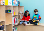 Photo: On White Wall Inc.Image description: Two children read books on a bench by a bookshelf.