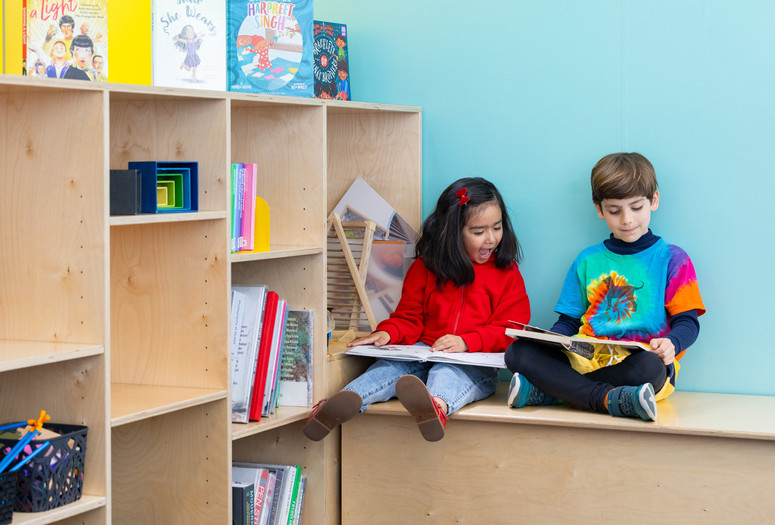 Photo: On White Wall Inc.Image description: Two children read books on a bench by a bookshelf.