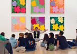 Photo: On White Wall Inc.Image description: A seated educator gestures with open arms while teaching families in front of six colorful square paintings of flowers.