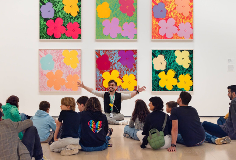 Photo: On White Wall Inc.Image description: A seated educator gestures with open arms while teaching families in front of six colorful square paintings of flowers.