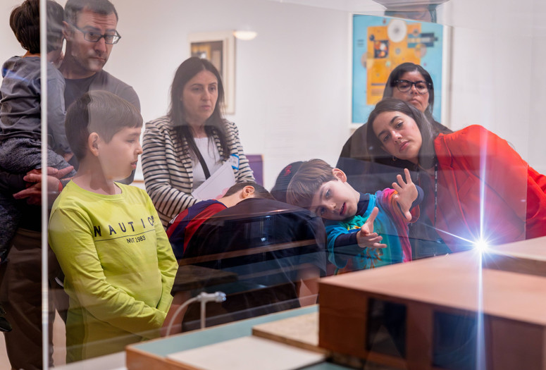 Photo: On White Wall, Inc.Image description: An educator and families view a work displayed behind glass together.