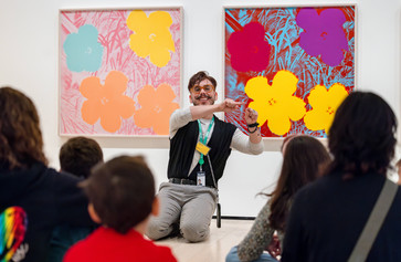 A smiling educator explains two works of art to seated families. Photo: On White Wall Inc.