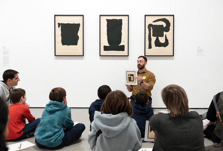 Photo: Martin SeckImage description: An educator holds up an image in front of three artworks while families observe.