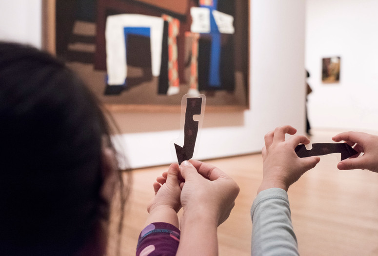 Photo: Martin SeckImage description: Children hold up laminated paper cutouts in front of an artwork.