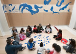 Photo: Martin SeckImage description: An educator leads a discussion with a group of families seated around blue cutout pieces
