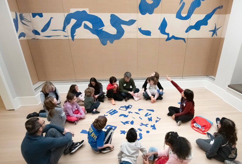 Photo: Martin SeckImage description: An educator leads a discussion with a group of families seated around blue cutout pieces