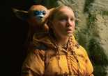 The Legend of Ochi. 2025. USA/Finland/UK. Directed by Isaiah Saxon. Courtesy of A24 Image description: A creature with pointy ears, red fur, a blue face, and large black eyes sits on the shoulders of a teenager with blonde braided hair and blue eyes wearing a yellow winter coat. Both stare with open mouths into the distance in front of a dirt wall.
