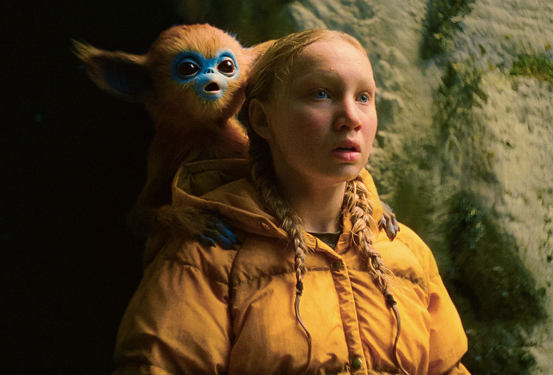 The Legend of Ochi. 2025. USA/Finland/UK. Directed by Isaiah Saxon. Courtesy of A24 Image description: A creature with pointy ears, red fur, a blue face, and large black eyes sits on the shoulders of a teenager with blonde braided hair and blue eyes wearing a yellow winter coat. Both stare with open mouths into the distance in front of a dirt wall.