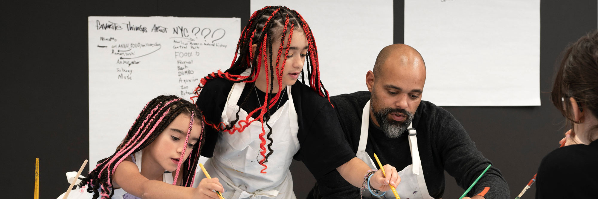 Photo: Martin SeckImage description: A caregiver and two children dip their paintbrushes into paint palettes and work on individual artworks