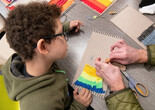An adult’s hands tie fabric onto a cardboard loom while a child looks on. Photo: Martin Seck