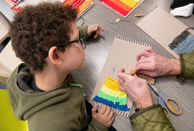 An adult’s hands tie fabric onto a cardboard loom while a child looks on. Photo: Martin Seck