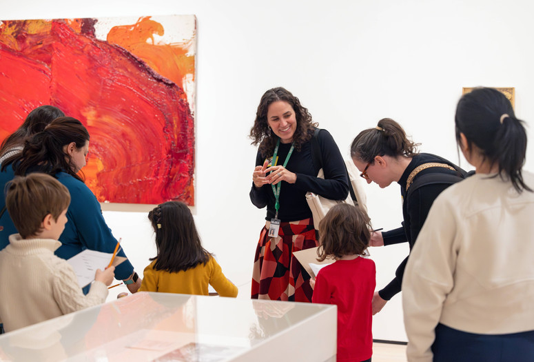An educator leads a gallery talk for a group of families in front of a bright red painting. Photo: On White Wall Inc.