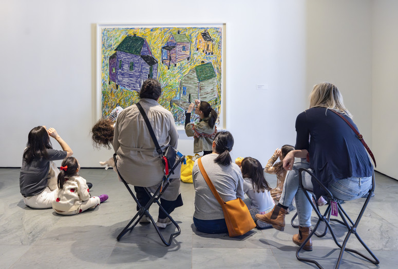 An educator looks up at an artwork depicting houses, and uses her hands as binoculars while she leads a group of families in a conversation. Photo courtesy On White Wall, Inc.