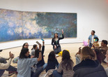 An educator kneels in front of an artwork, holding a pink color card, and leads a discussion about the art with a group of families. Photo: On White Wall, Inc.